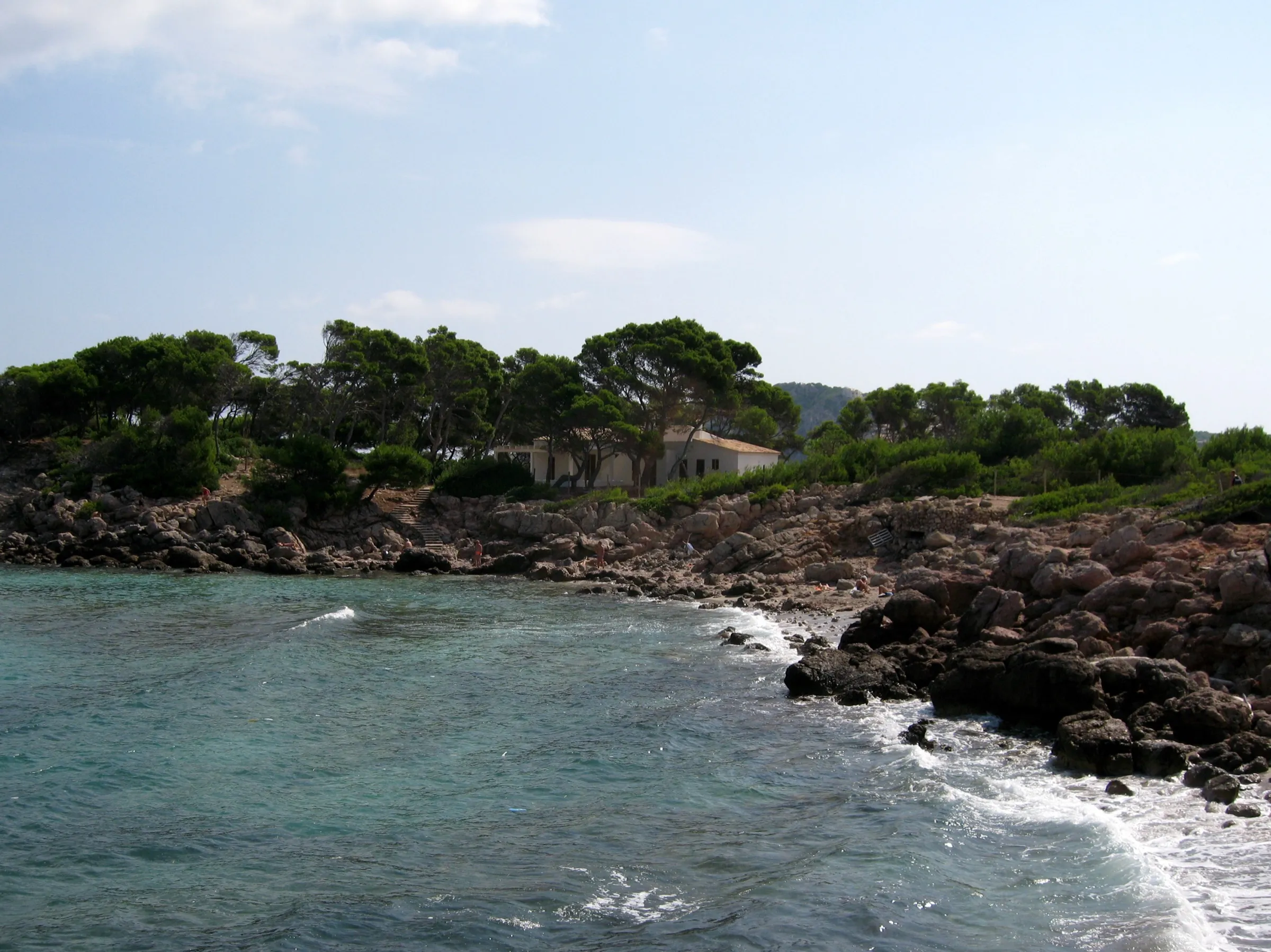 El lado sudeste de Cala Moltó, con rocas oscuras en la orilla, agua clara y pinos detrás