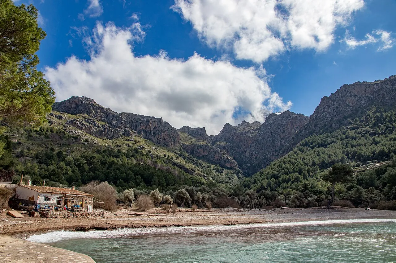 La orilla de Cala Tuent con una caseta a la izquierda y las paredes de roca de la Serra de Tramuntana al fondo
