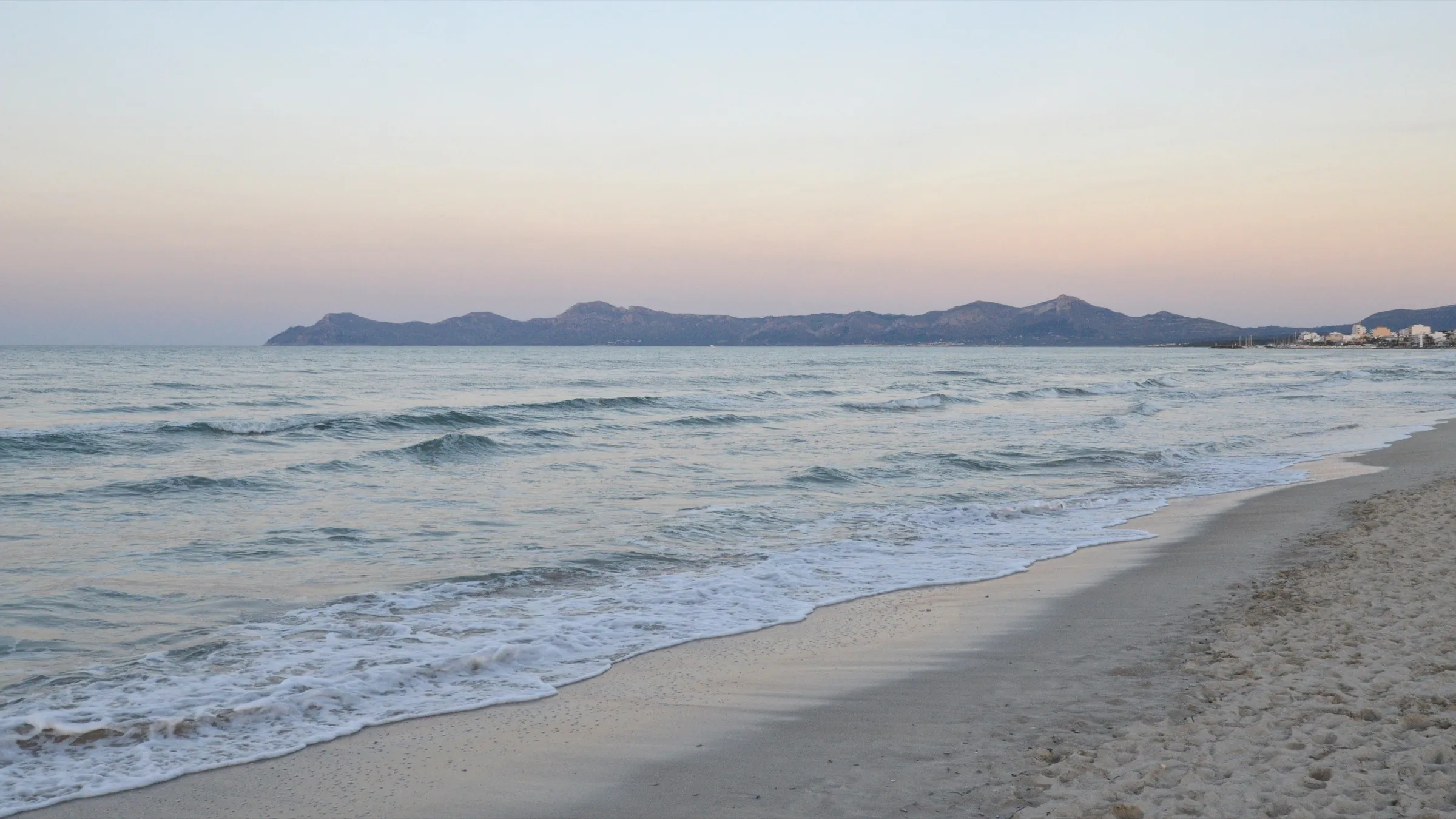 Orilla de Es Comú al atardecer, con olas suaves y las montañas de Alcúdia al otro lado de la bahía