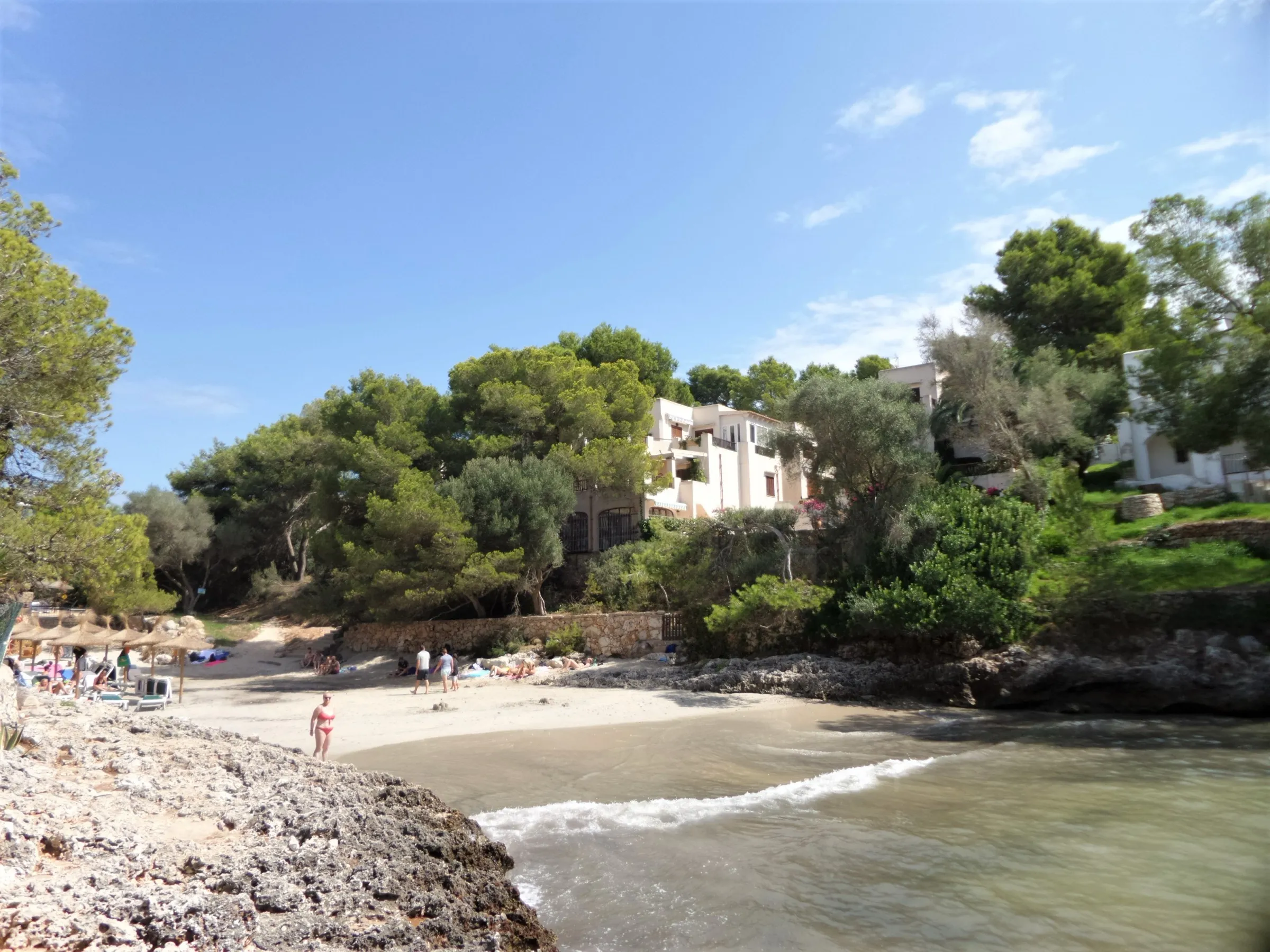Der Sand der Cala Serena von den Felsen aus, mit Badegästen und einem weißen Haus zwischen den Pinien