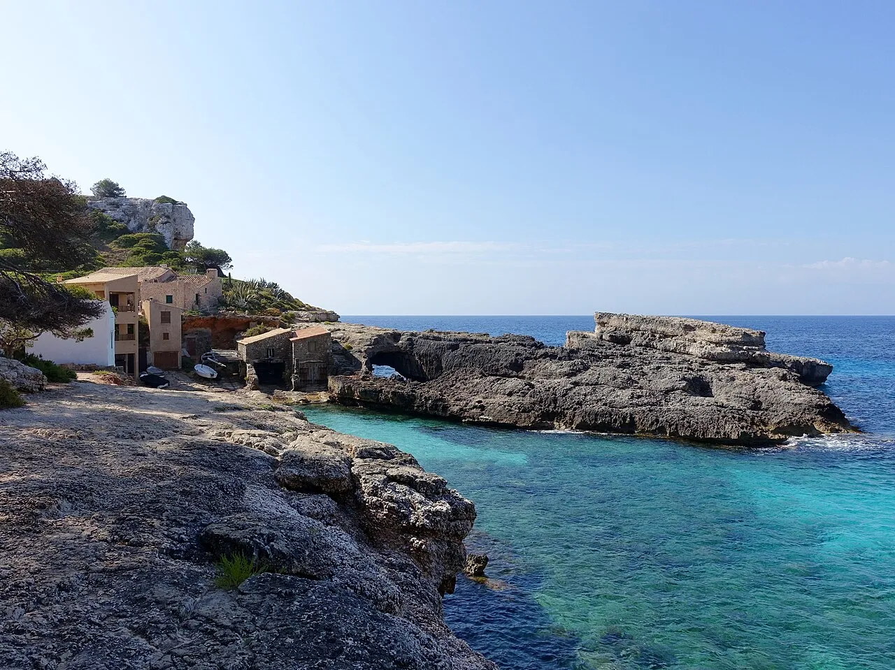 Plataforma de roca con un arco natural en la boca de Cala s'Almunia, junto a las casetas y el mar abierto