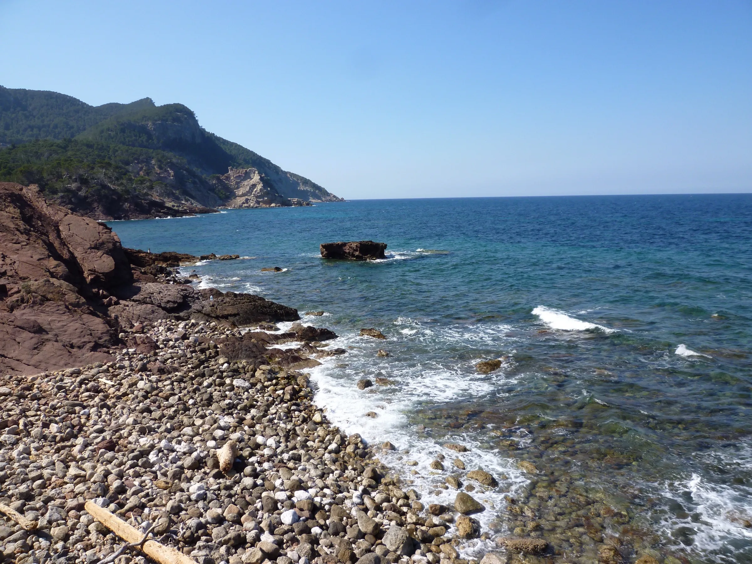 Orilla de cantos redondos junto a la roca rojiza del Port des Canonge, con el mar y la sierra al fondo