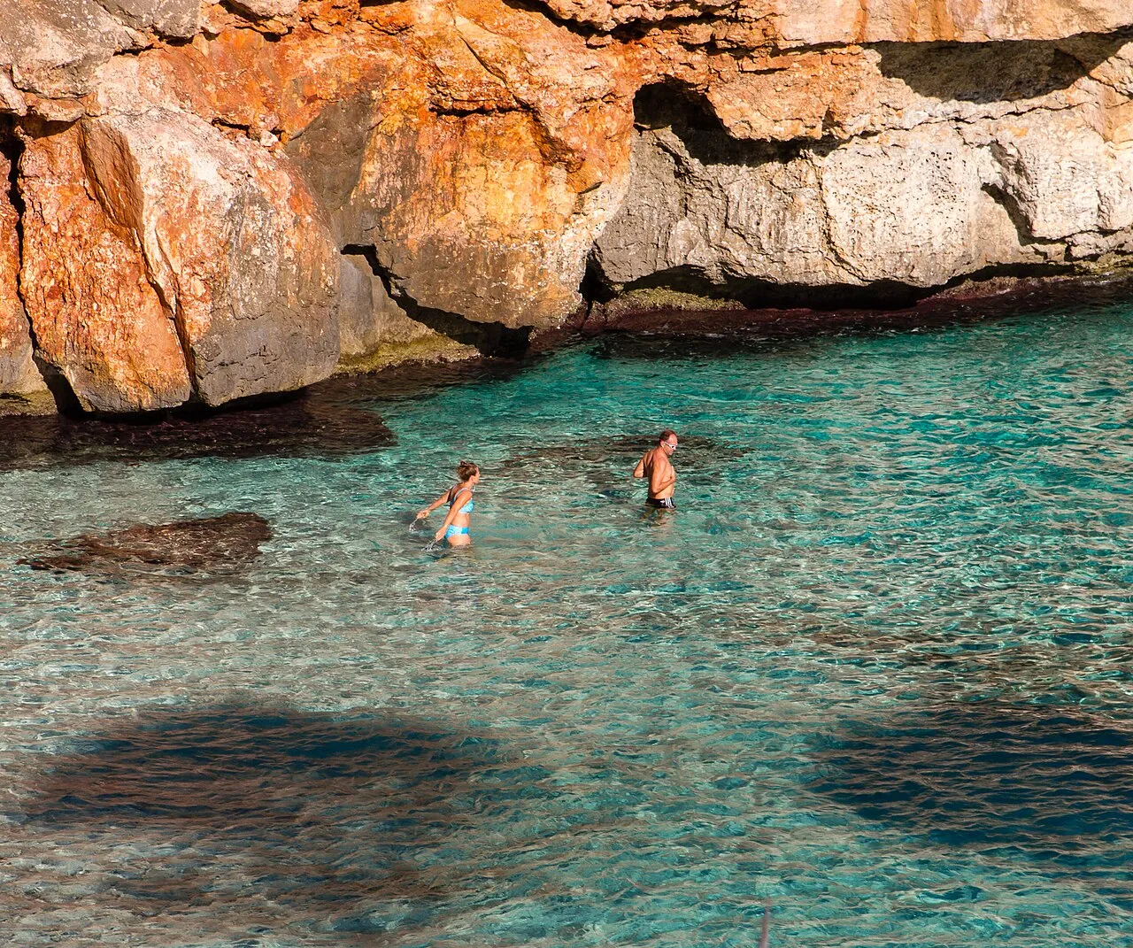 Dos bañistas en el agua transparente del Caló des Moro, bajo una pared de roca anaranjada