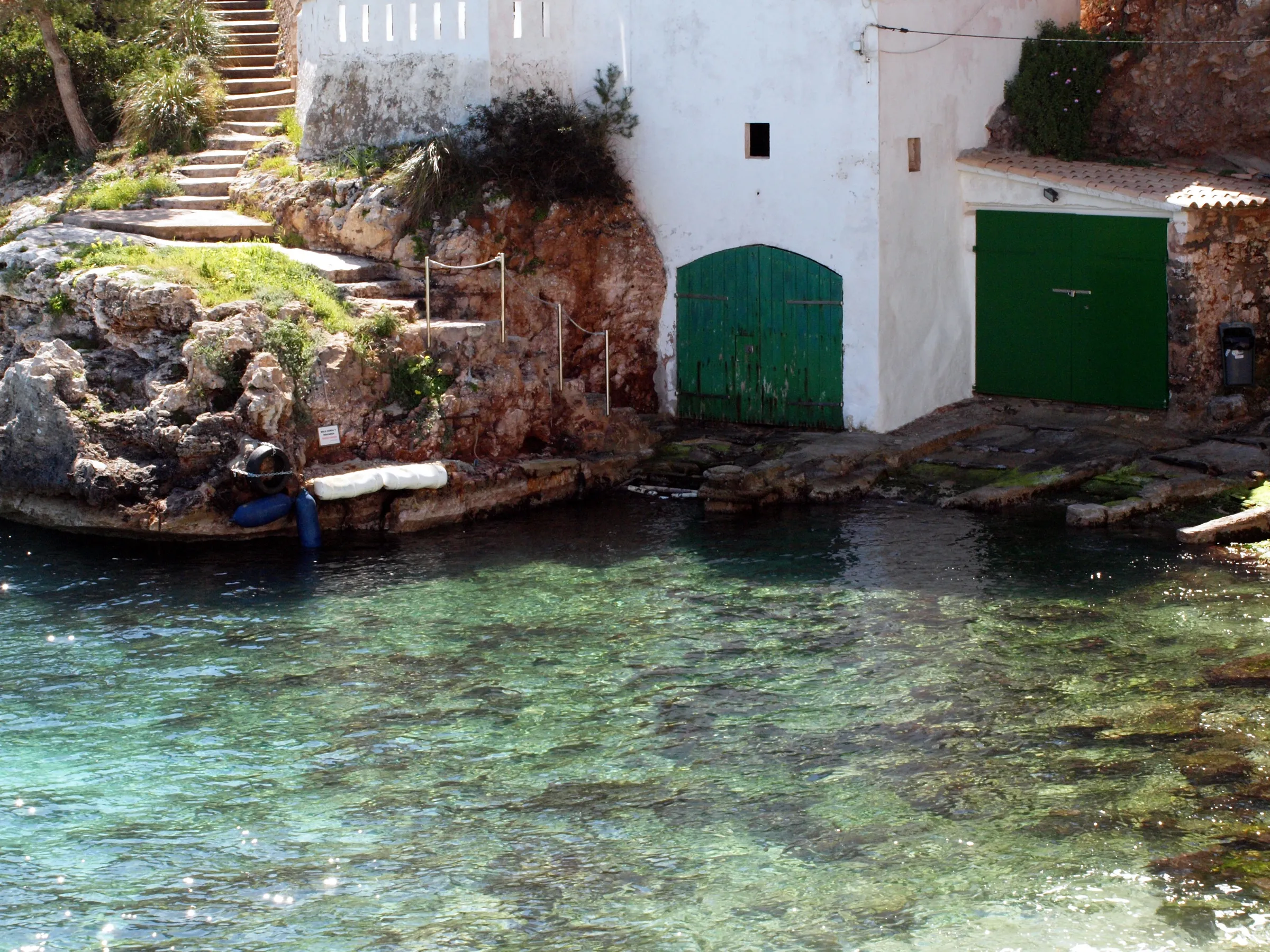 Casetas de barcas con puertas verdes y escalones de piedra junto al agua clara de Cala Santanyí