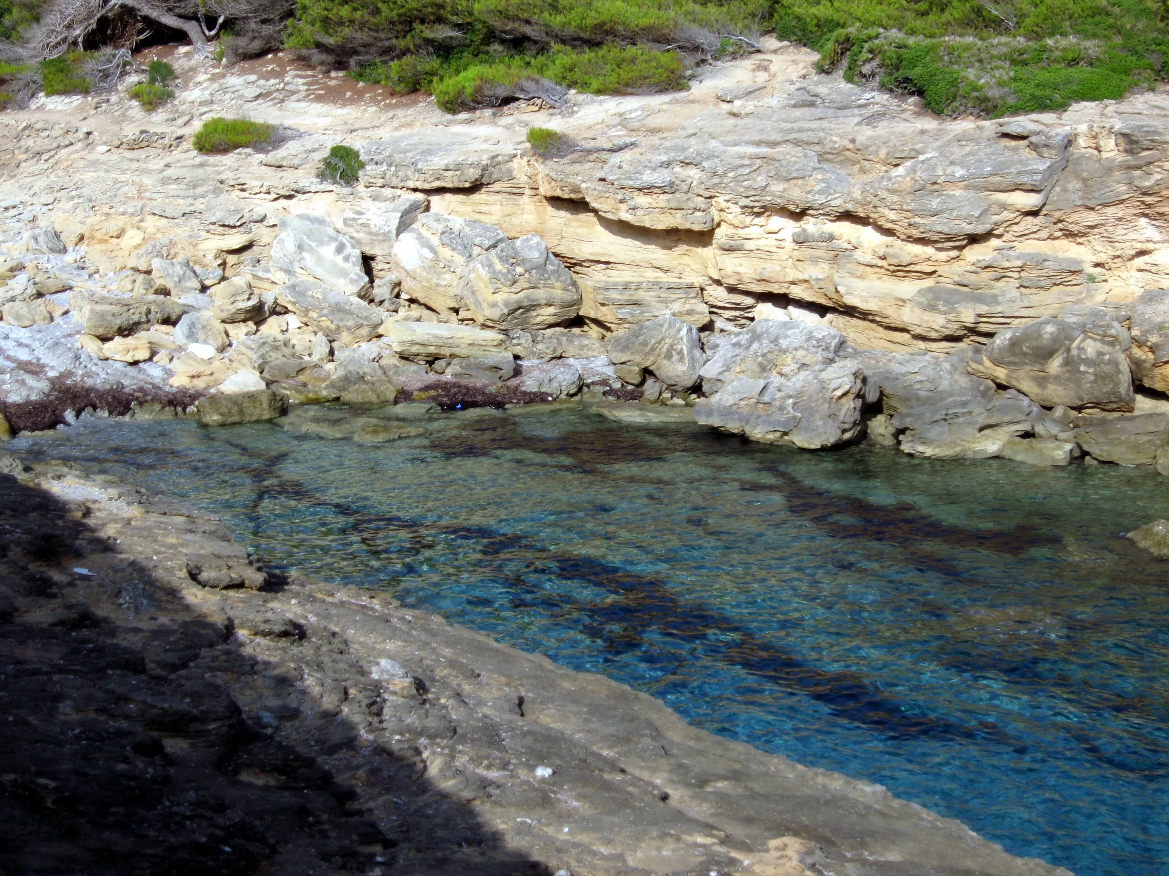 Agua transparente sobre el fondo de roca de Cala Estreta, bajo capas de piedra caliza