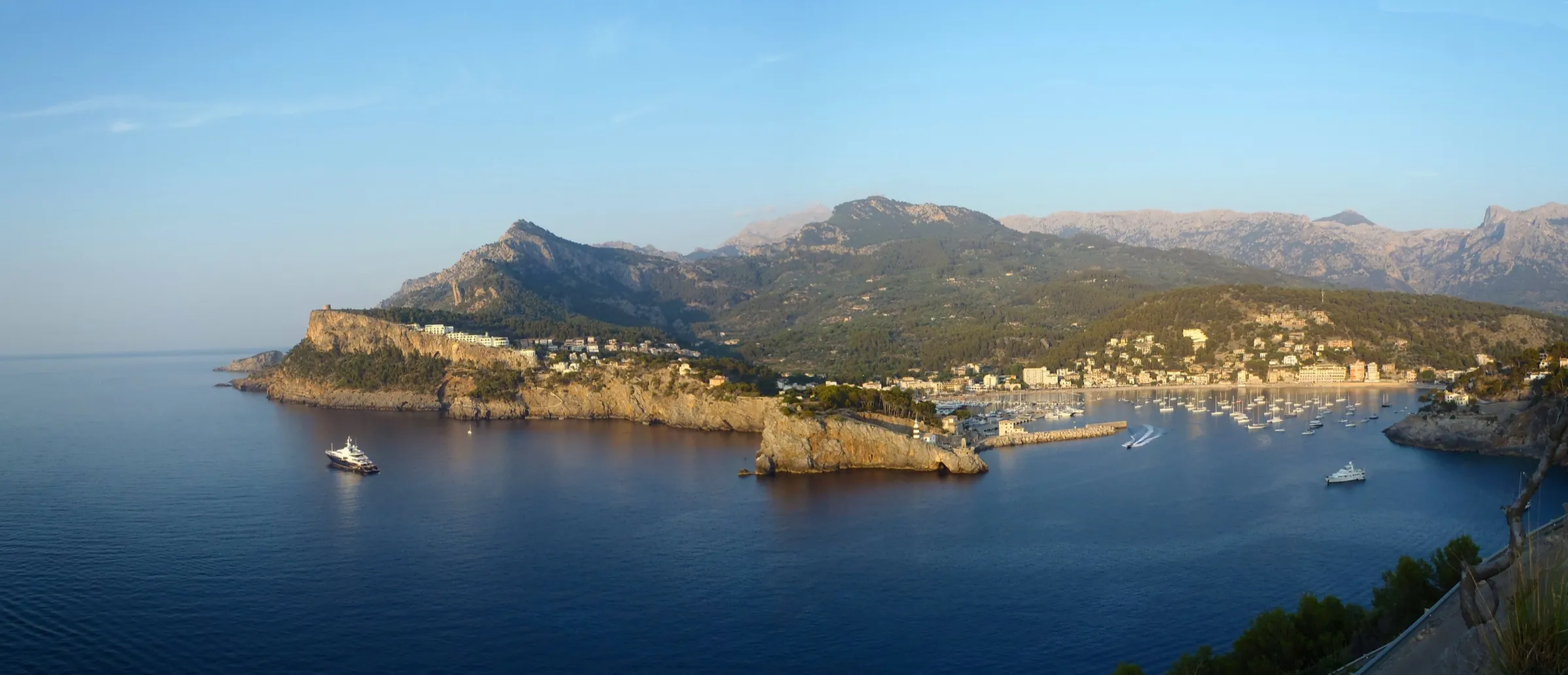 La bahía del Port de Sóller vista desde lo alto al atardecer, con el faro, el puerto, las playas del fondo y la Serra de Tramuntana detrás
