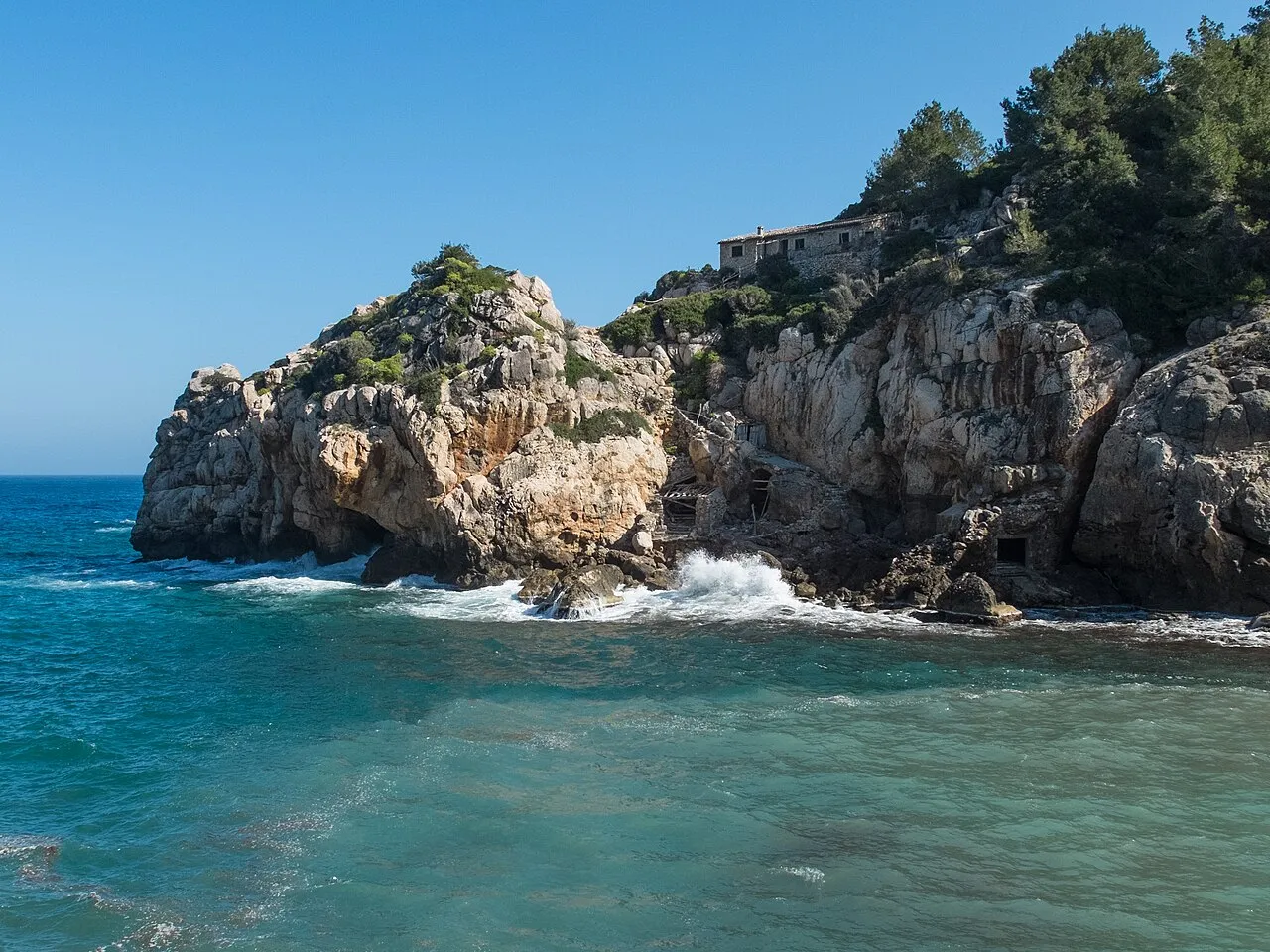 Promontorio de roca con pinos en la boca de Cala Deià y el mar rompiendo a sus pies