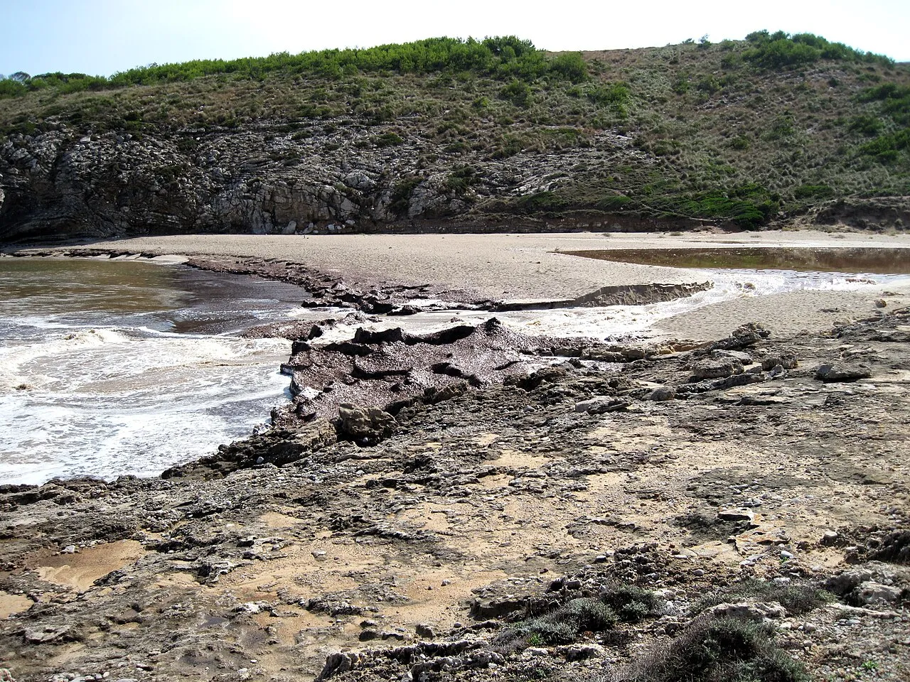 Vista de la cala desde la costa rocosa, con el agua turquesa y la arena al fondo del valle