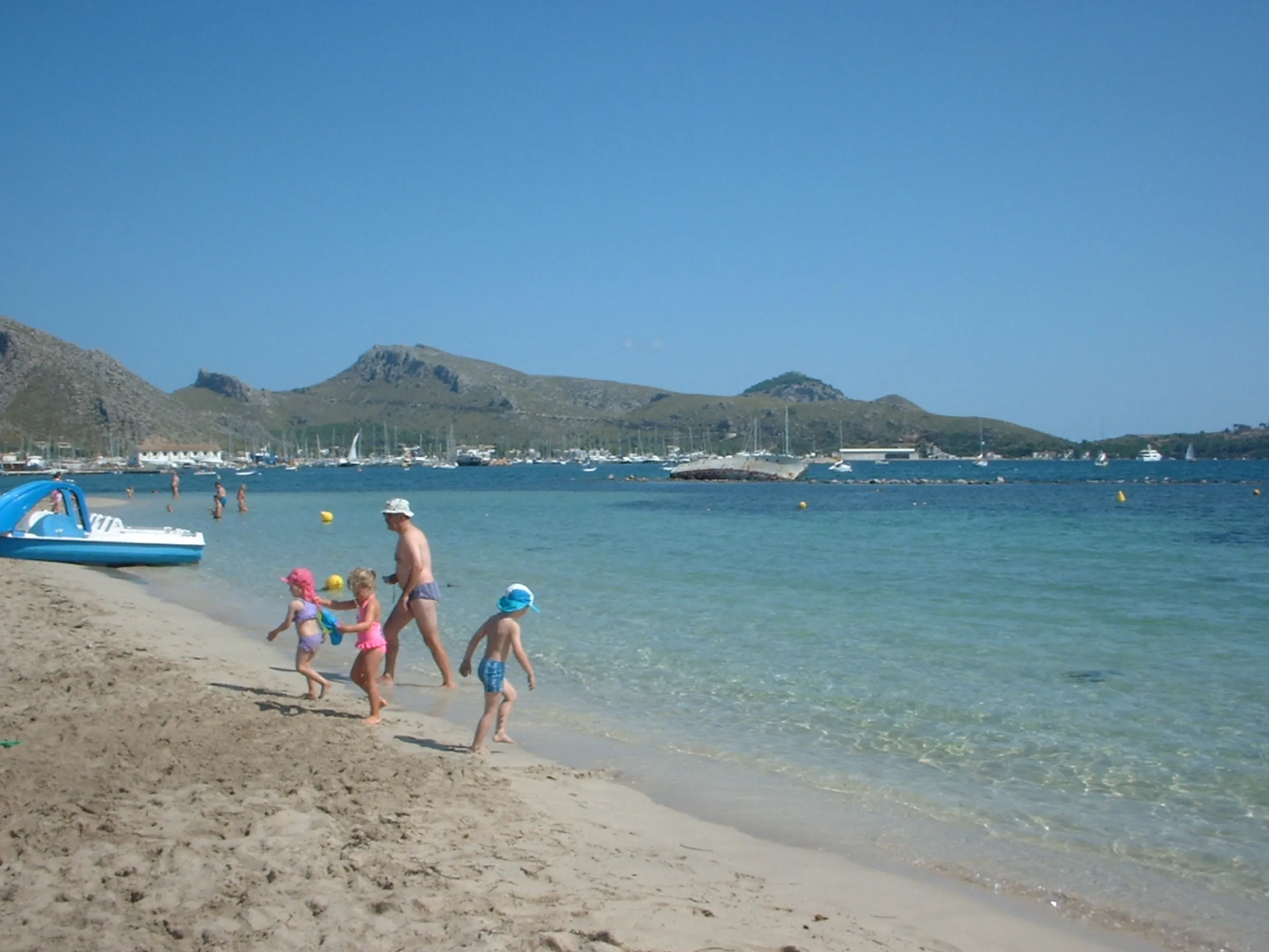 A family playing at the water's edge in Llenaire, with clear water and boats anchored in the bay