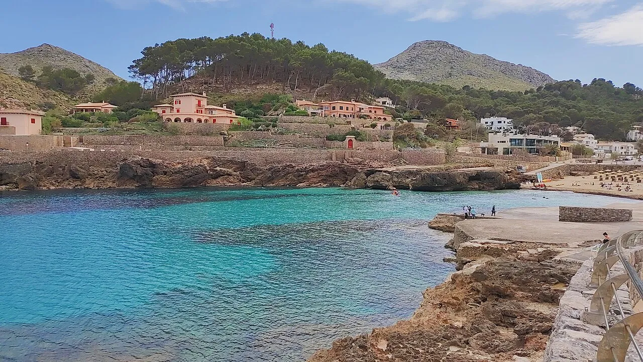 Cala Molins, con el agua turquesa entre rocas bajas y las casas del pueblo en la ladera