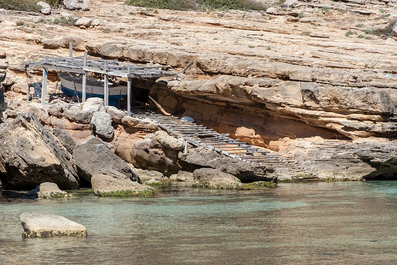 Escar con rampa de madera bajo una cornisa de roca, junto al agua transparente de Cala Figuera