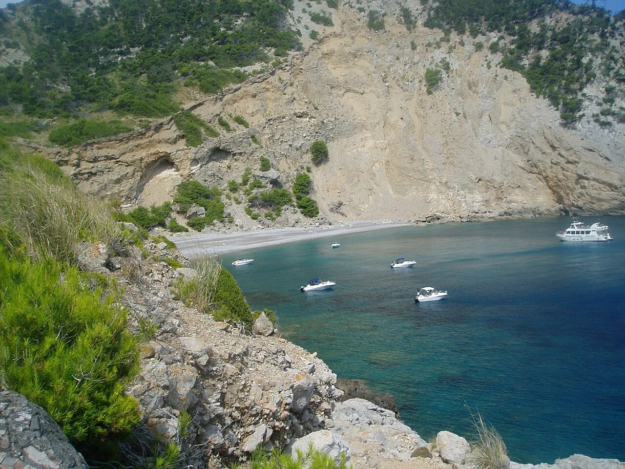 Vista desde lo alto de la cala de es Coll Baix, con barcas en el agua y el acantilado detrás de la playa