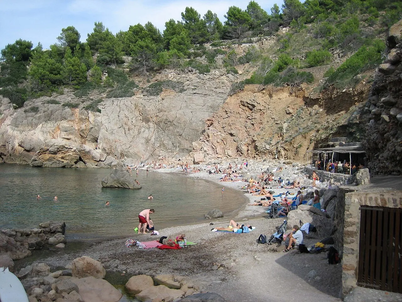 Bañistas en la orilla de cantos de Cala Deià, bajo el acantilado y el pinar