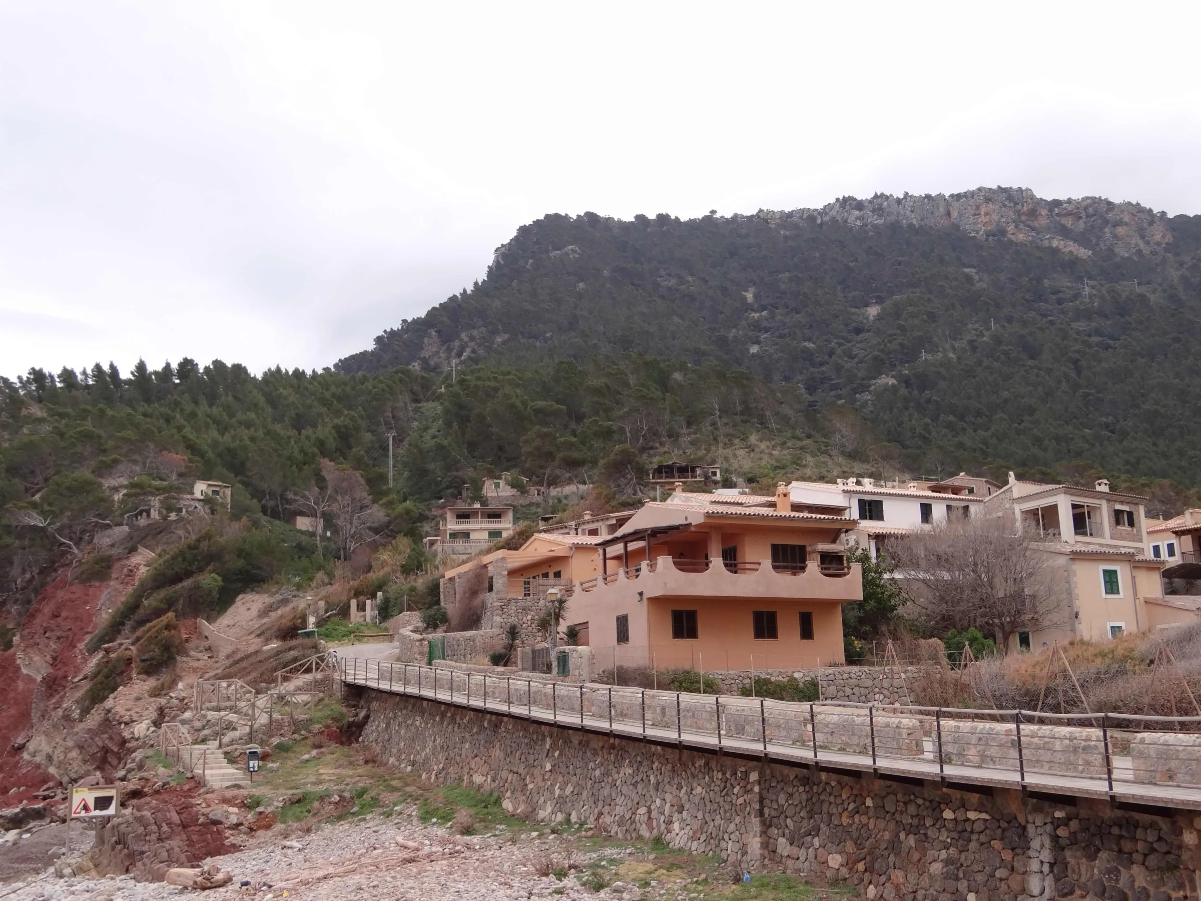 La orilla de cantos del Port de Valldemossa, con un muro de piedra y casas entre el pinar