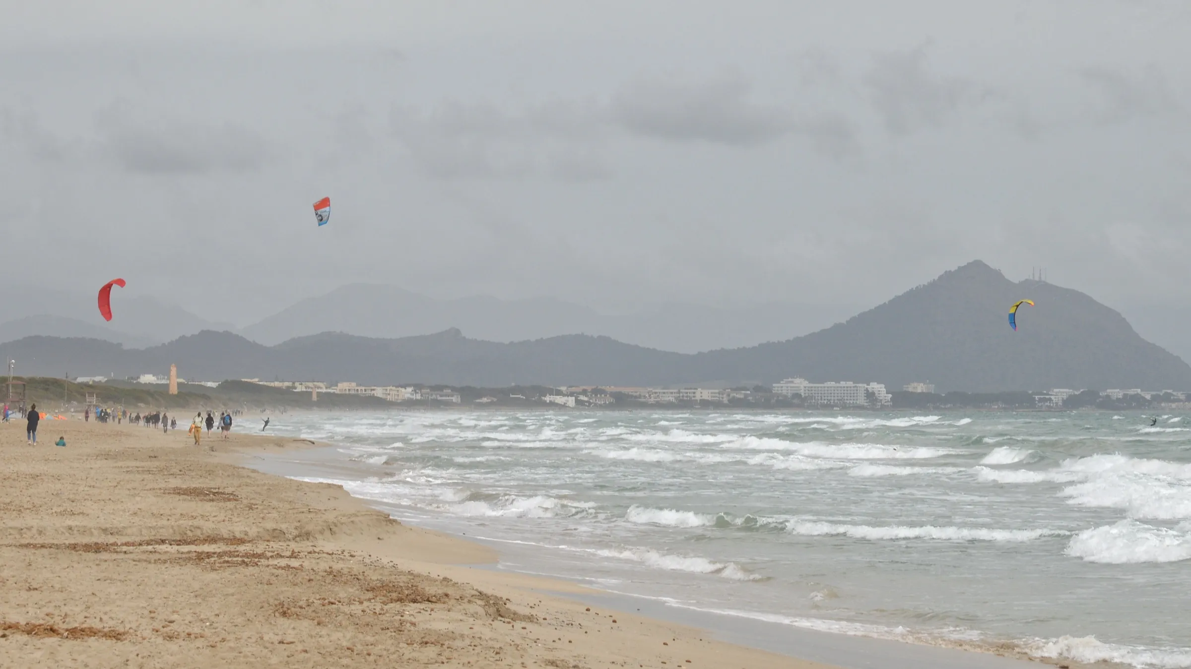 Waves at the water's edge in Can Picafort on a windy day, with kitesurfing kites and the Alcúdia mountains in the distance