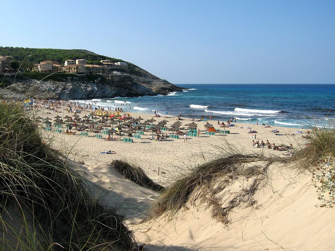 Sombrillas y hamacas en la arena de Cala Mesquida, vistas desde la duna, con olas en la orilla