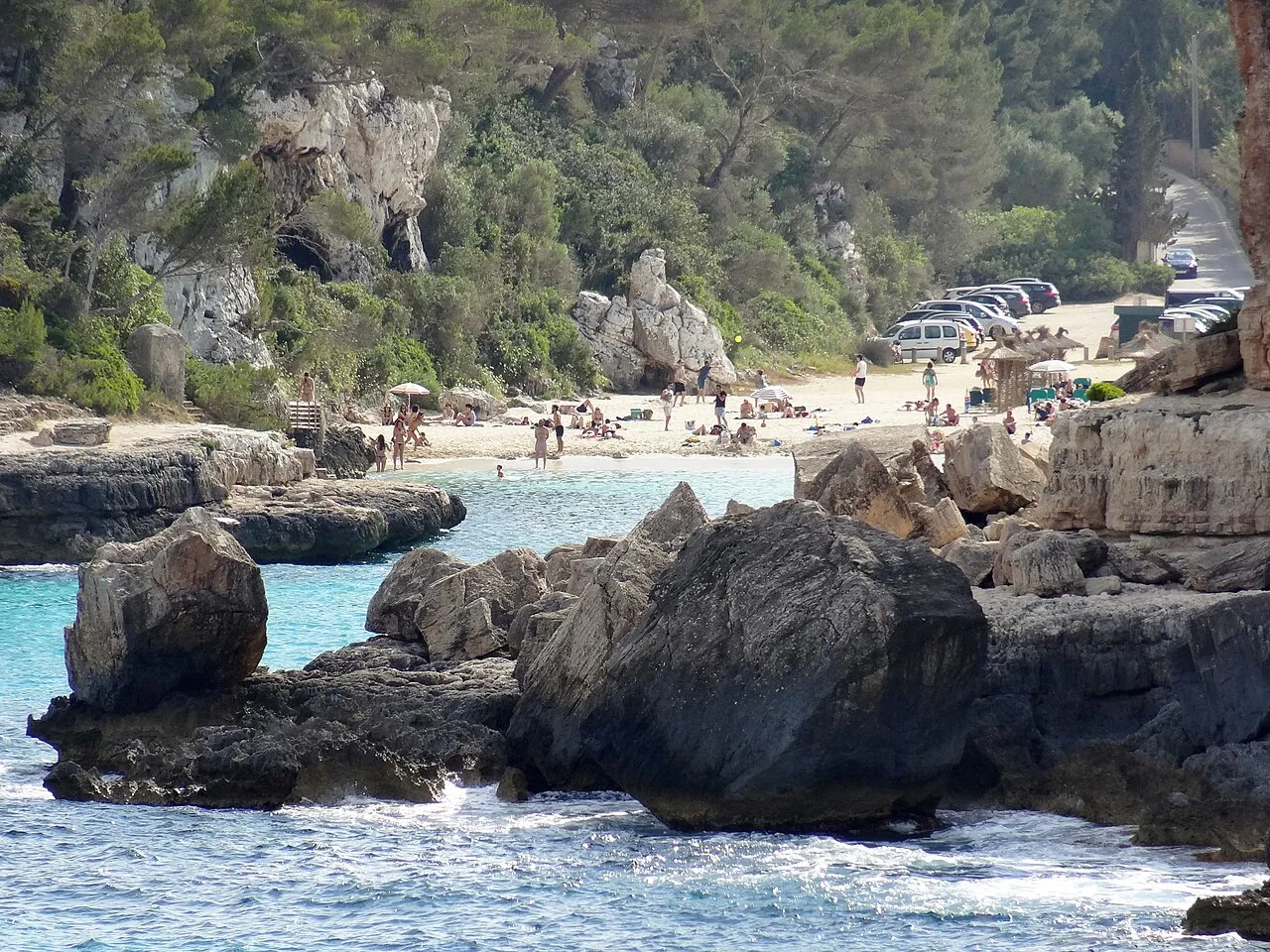 Rocas junto al agua en Cala Llombards, con la playa y los coches aparcados al fondo