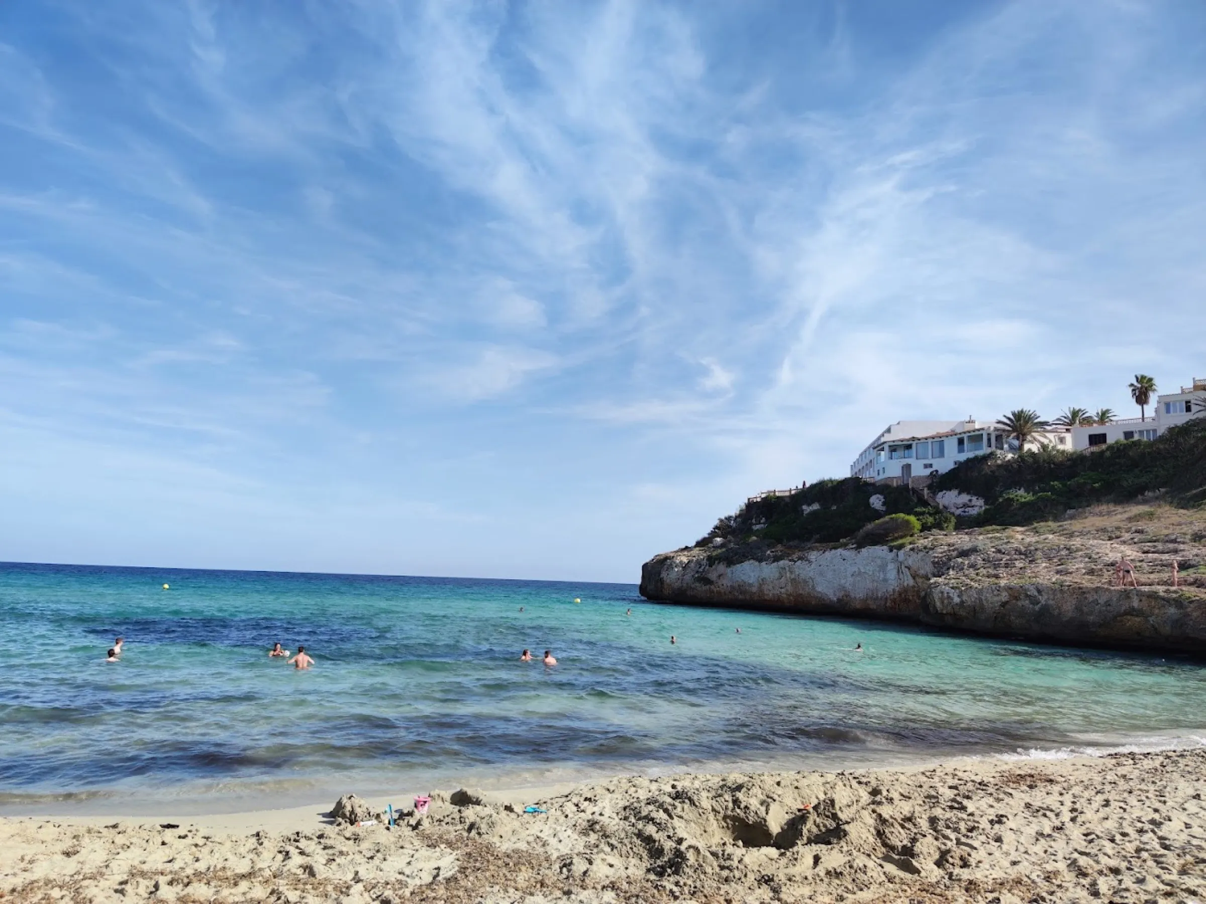 Agua turquesa en la orilla de Cala Domingos, con la punta de roca y casas blancas al fondo