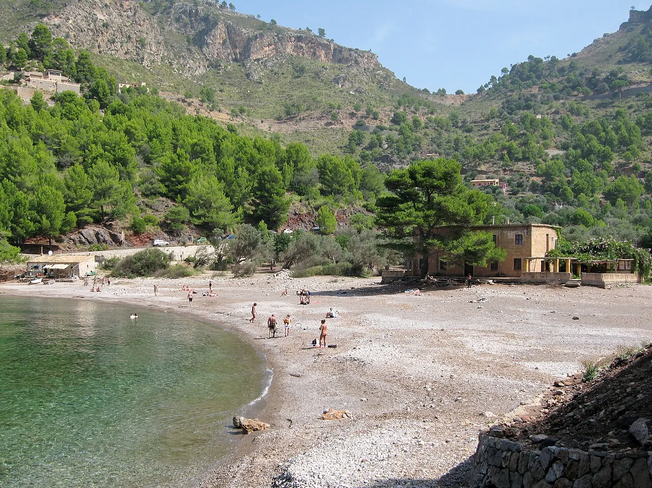 La playa de cantos de Cala Tuent con algunos bañistas, una casa entre pinos y las laderas del valle detrás